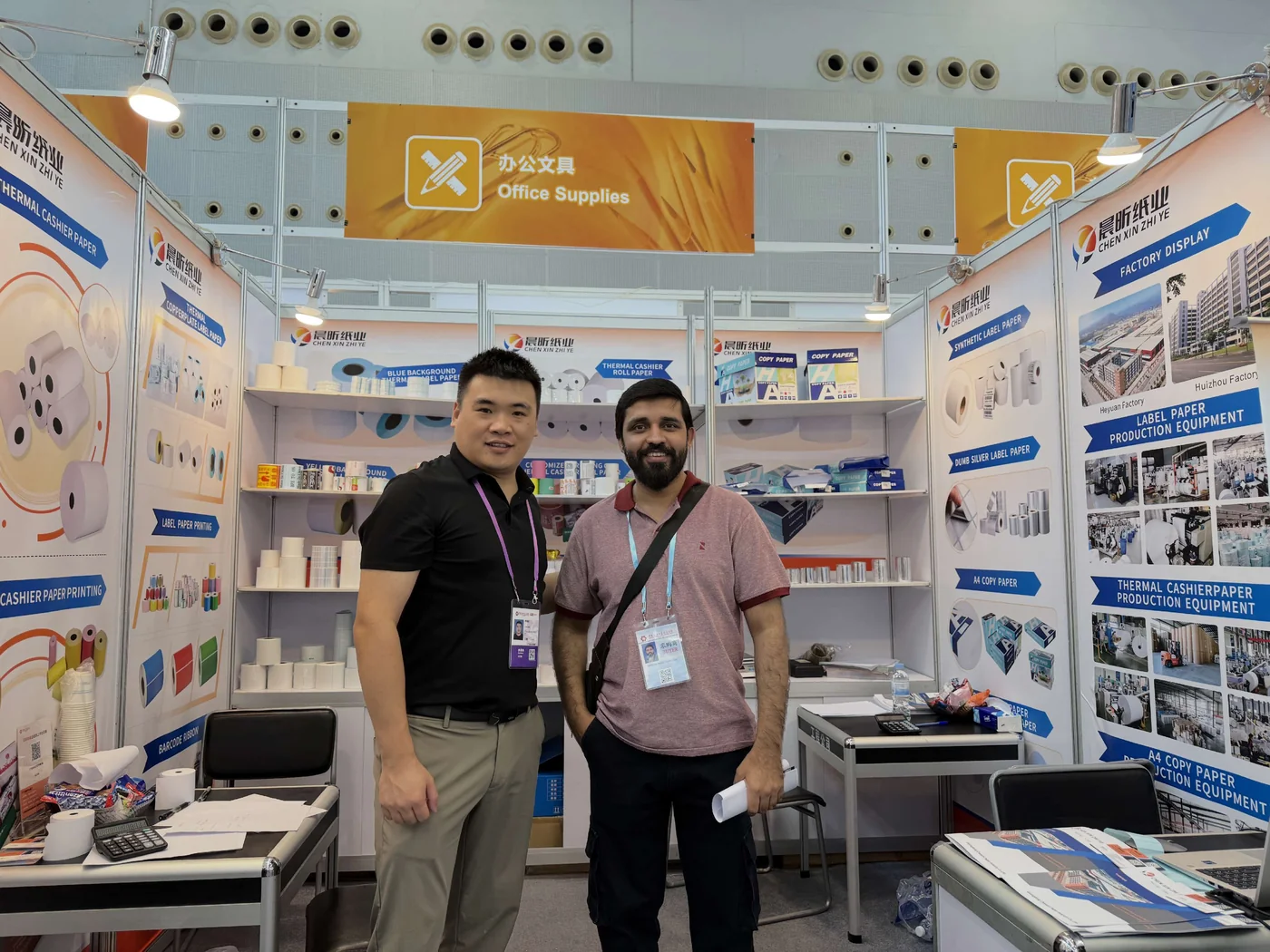 Two people posing together in front of paper-product displays at the exhibition